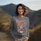 A woman smiles, wearing a grey sweatshirt with "Baddest Witch in Town" and a witch graphic.