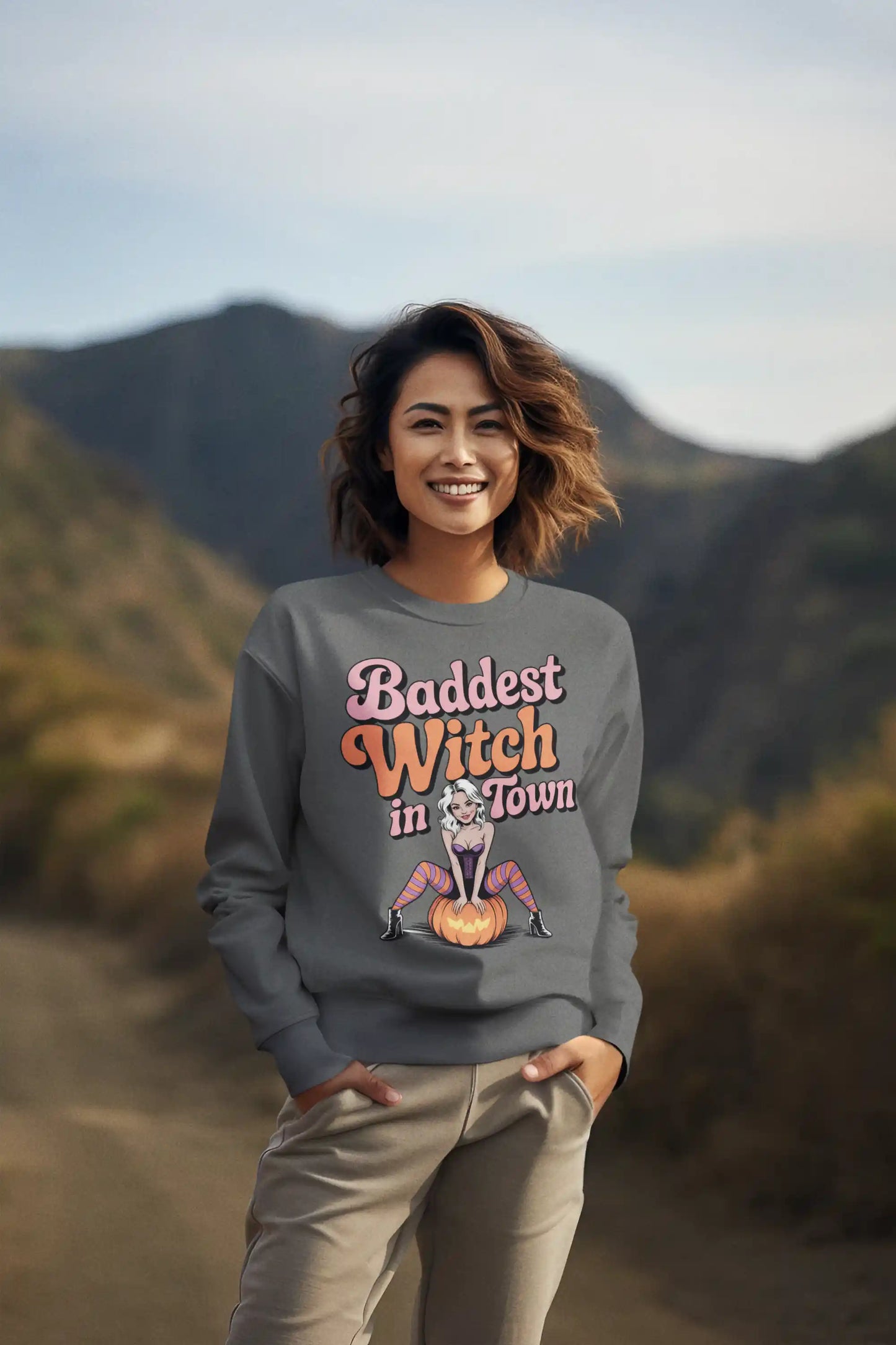 A woman smiles, wearing a grey sweatshirt with "Baddest Witch in Town" and a witch graphic.