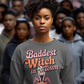 A woman wears a dark gray sweatshirt with "Baddest Witch in Town" and a witch on a pumpkin.