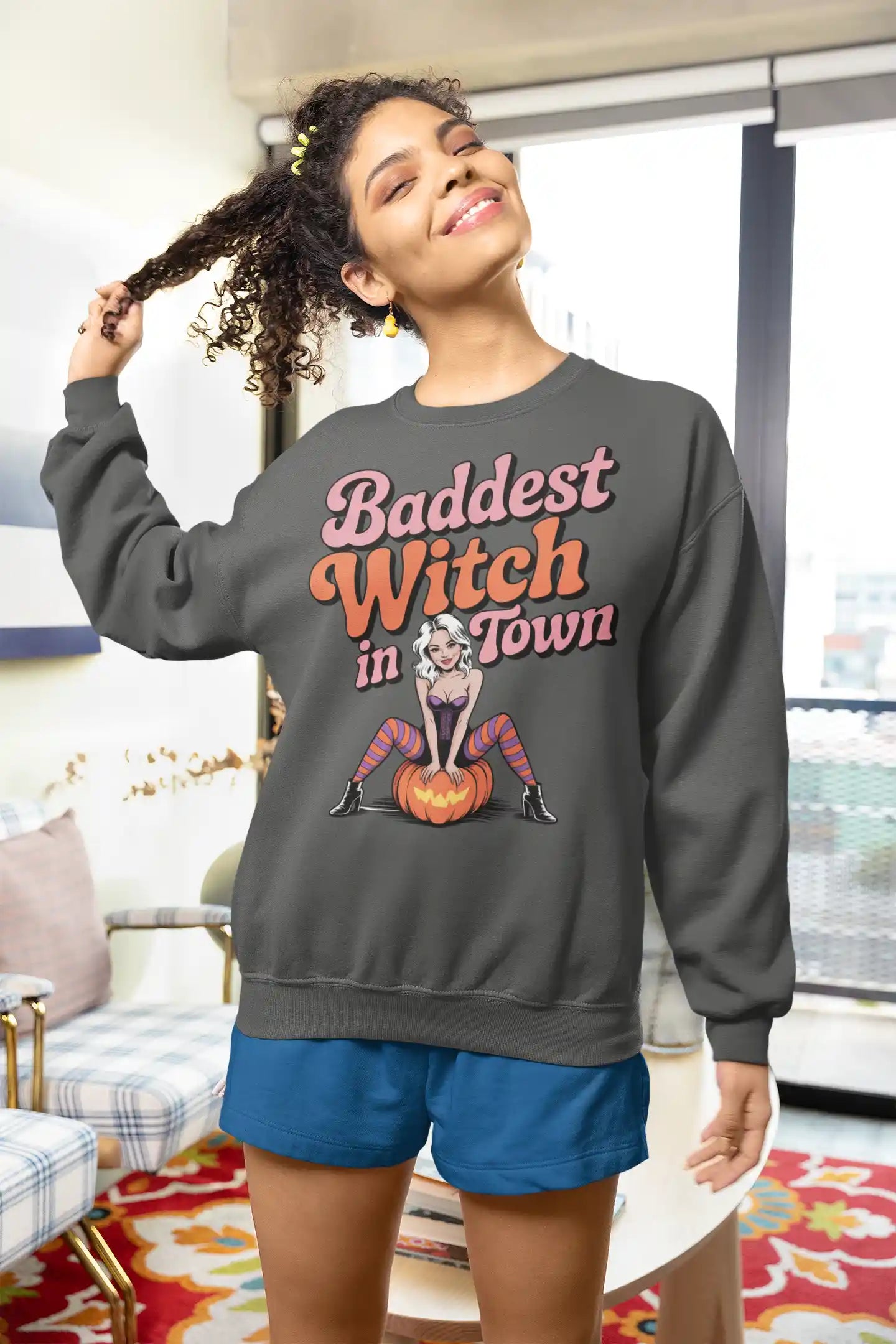 A woman wears a dark gray sweatshirt with "Baddest Witch in Town" and a witch on a pumpkin.