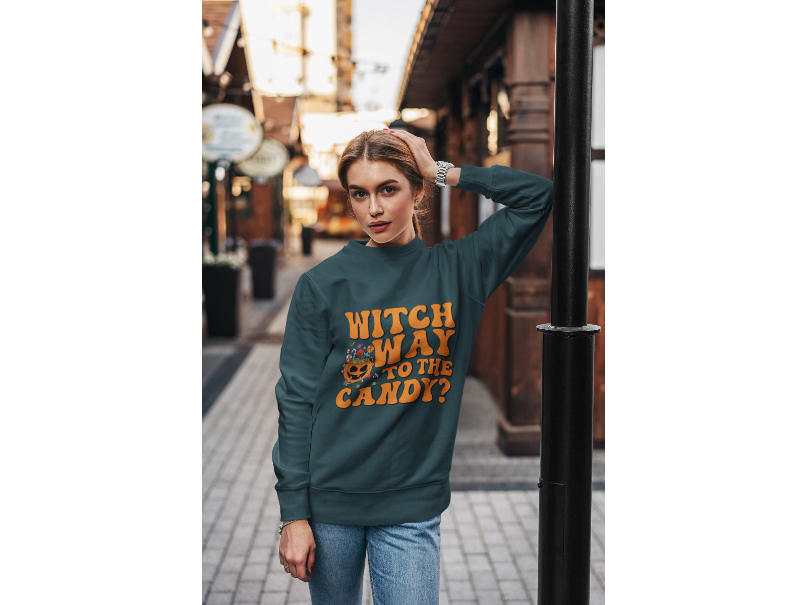A woman wears a dark green sweatshirt with "Witch Way to the Candy?" in orange text.