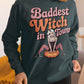 A woman wears a dark green sweatshirt with "Baddest Witch in Town" and a witch graphic.