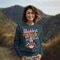 A woman in a dark teal "Baddest Witch in Town" sweatshirt stands outdoors.