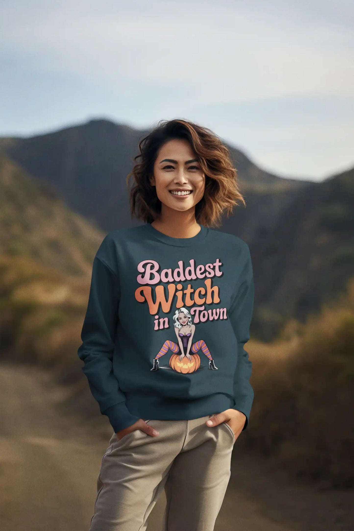 A woman in a dark teal "Baddest Witch in Town" sweatshirt stands outdoors.