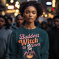 A woman wears a dark green sweatshirt with "Baddest Witch in Town" and a witch graphic.
