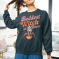 A woman wears a dark sweatshirt with "Baddest Witch in Town" and a witch on a pumpkin.