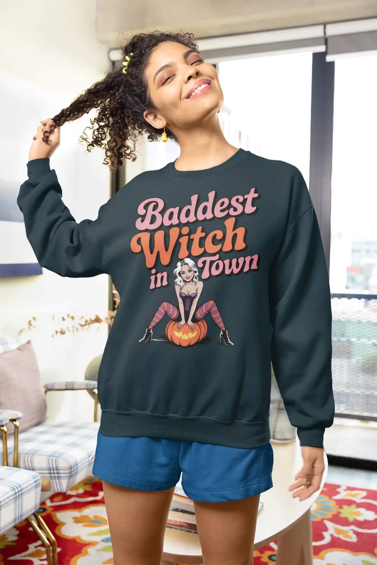 A woman wears a dark sweatshirt with "Baddest Witch in Town" and a witch on a pumpkin.