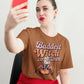 A woman in a brown "Baddest Witch in Town" t-shirt takes a selfie.