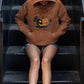 A person wearing a brown Halloween Town hoodie with a pumpkin graphic sits on metal stairs.