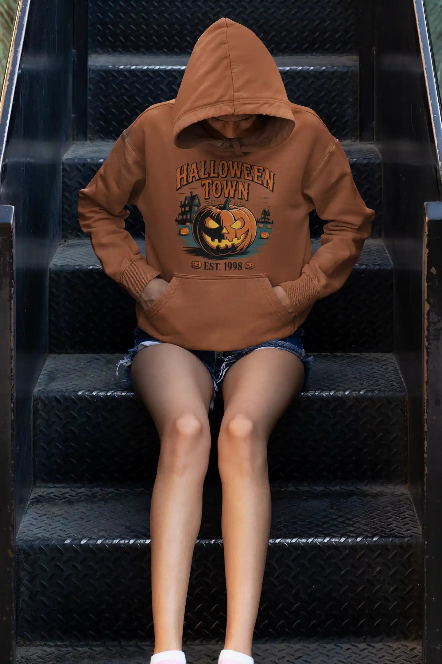 A person wearing a brown Halloween Town hoodie with a pumpkin graphic sits on metal stairs.