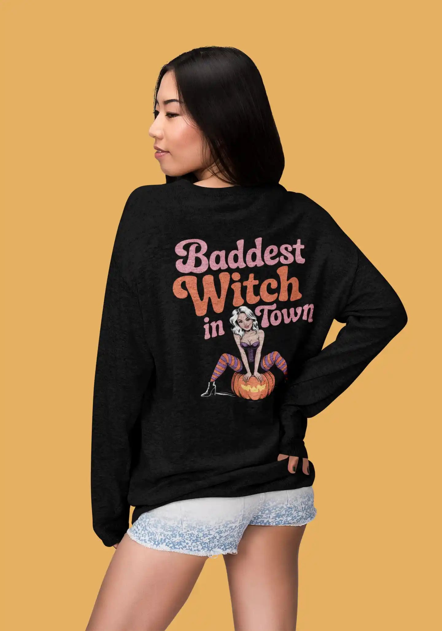 A woman models a black sweatshirt with "Baddest Witch in Town" and a witch graphic.
