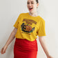 A woman smiles wearing a yellow Halloween Town t-shirt with a pumpkin graphic.