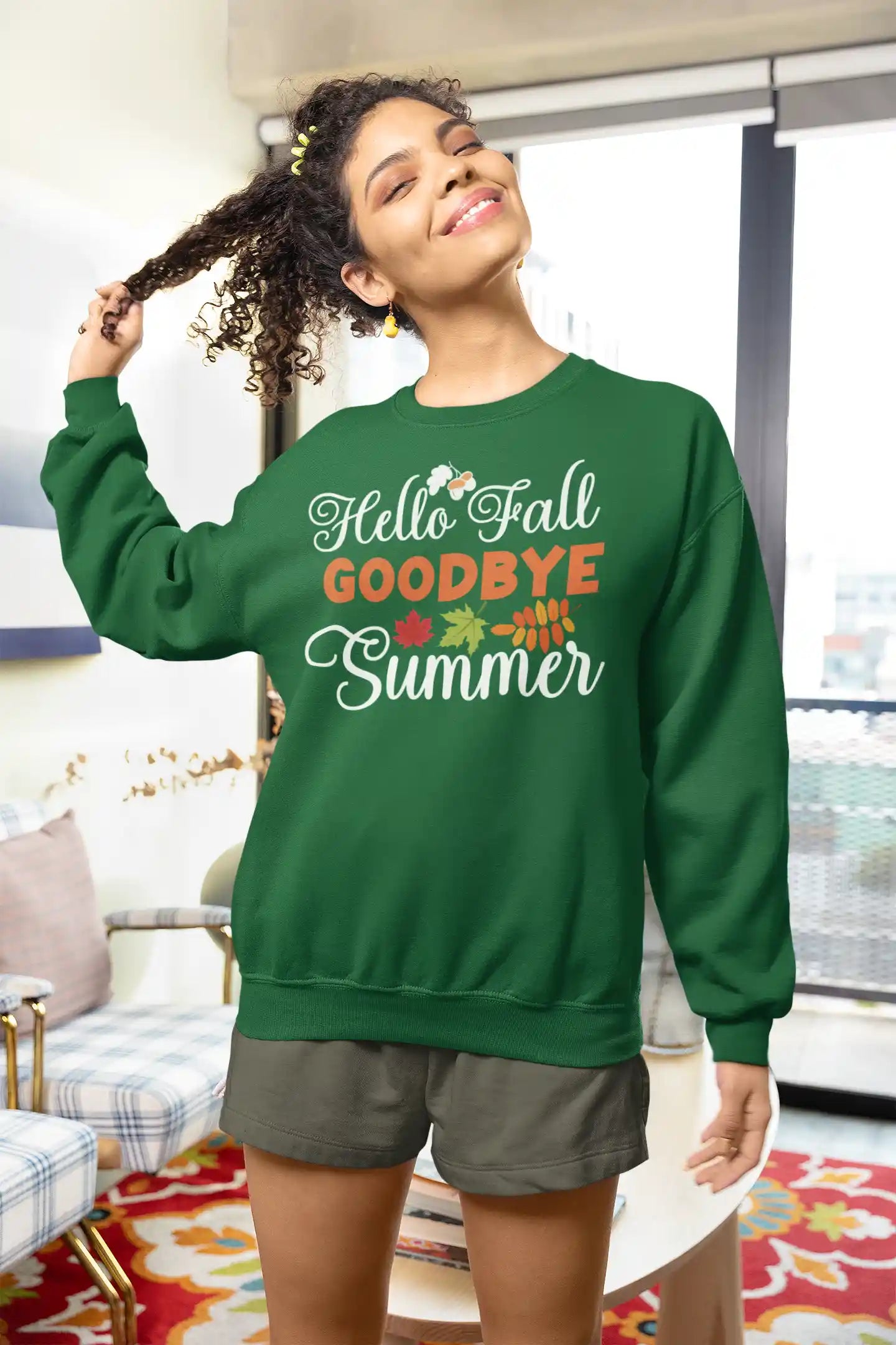 A woman in a green "Hello Fall Goodbye Summer" sweatshirt smiles, holding her curly hair.