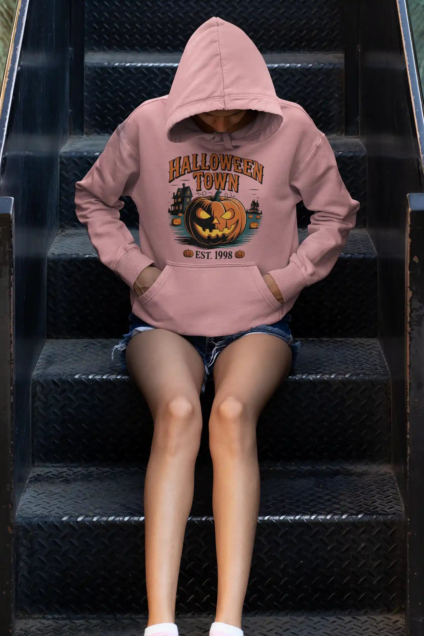 A person wearing a pink Halloween Town hoodie with a pumpkin graphic sits on metal stairs.