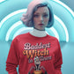 A woman wears a red sweatshirt with "Baddest Witch in Town" and a witch graphic.