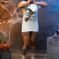 A woman in a white "Tonight We Fly" witch broom t-shirt stands in a Halloween setting.