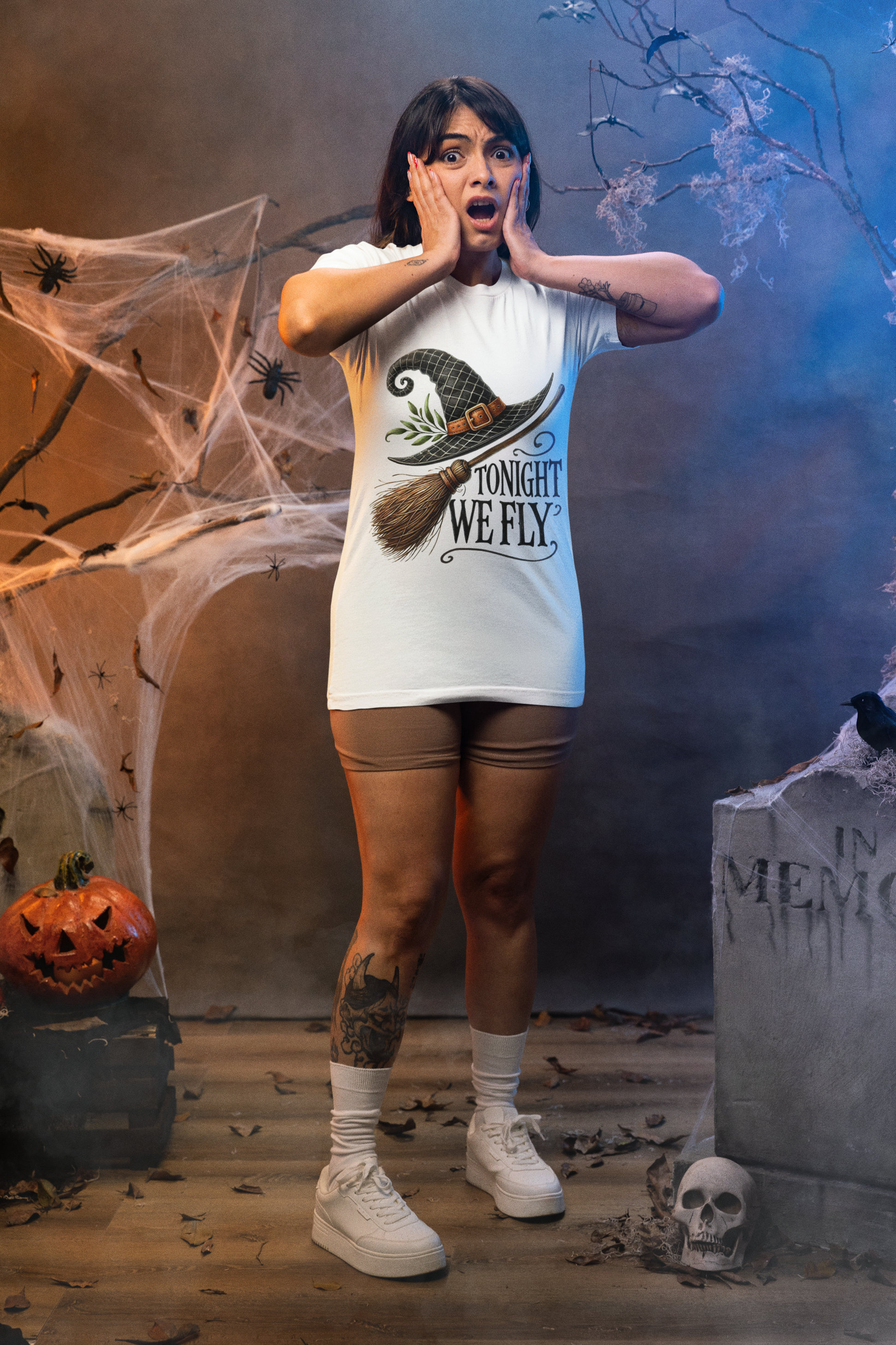 A woman in a white "Tonight We Fly" witch broom t-shirt stands in a Halloween setting.