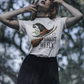 A woman wears a white t-shirt with a witch hat, broom, and "Tonight We Fly" graphic.