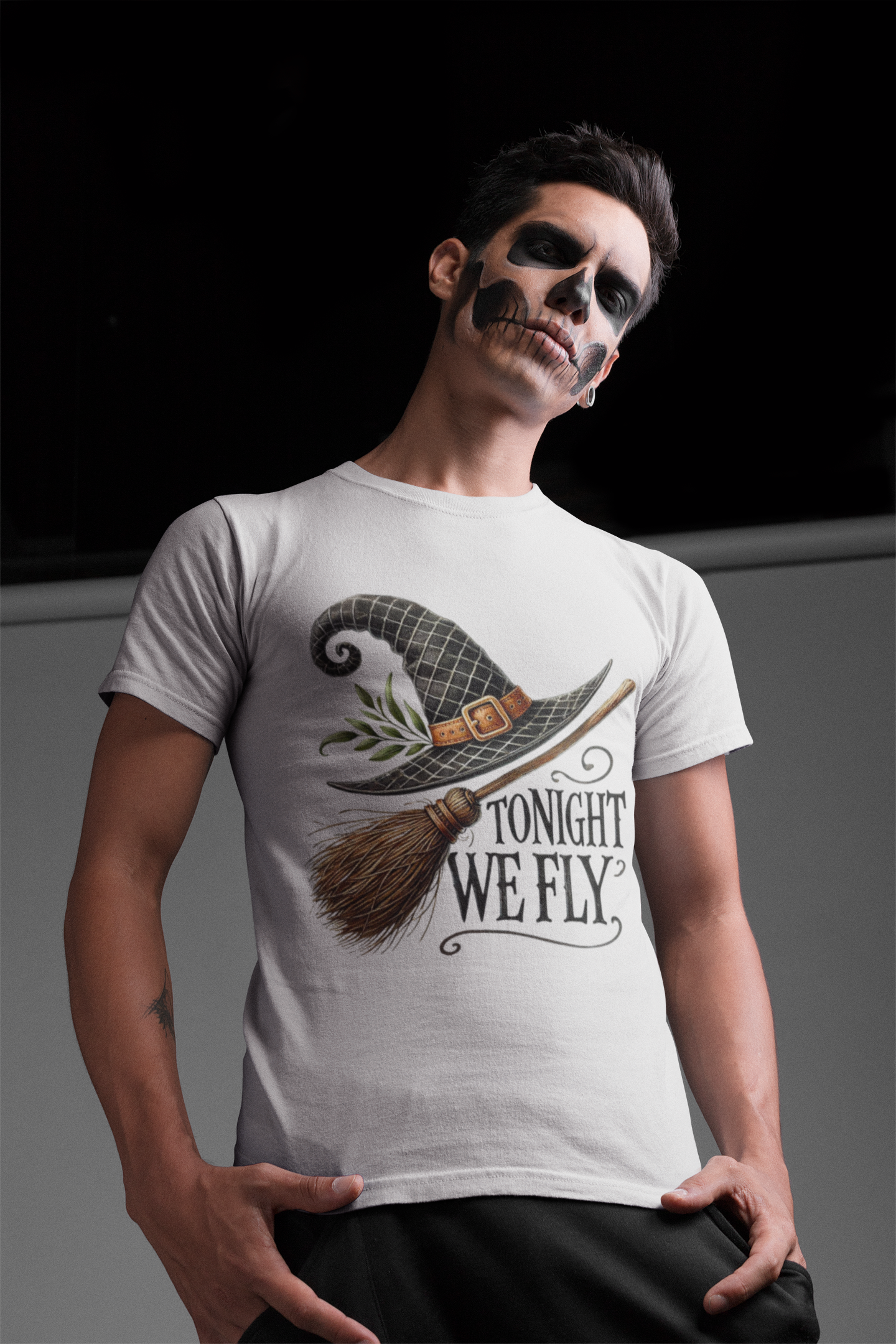 A man with skull makeup wears a white t-shirt with a witch hat and broom graphic.