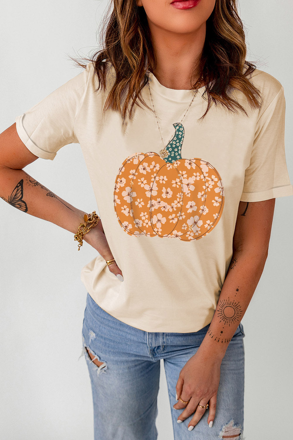 A woman wears a cream t-shirt with a floral pumpkin graphic and distressed jeans.