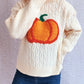 A cream cable-knit turtleneck sweater with an orange pumpkin applique.