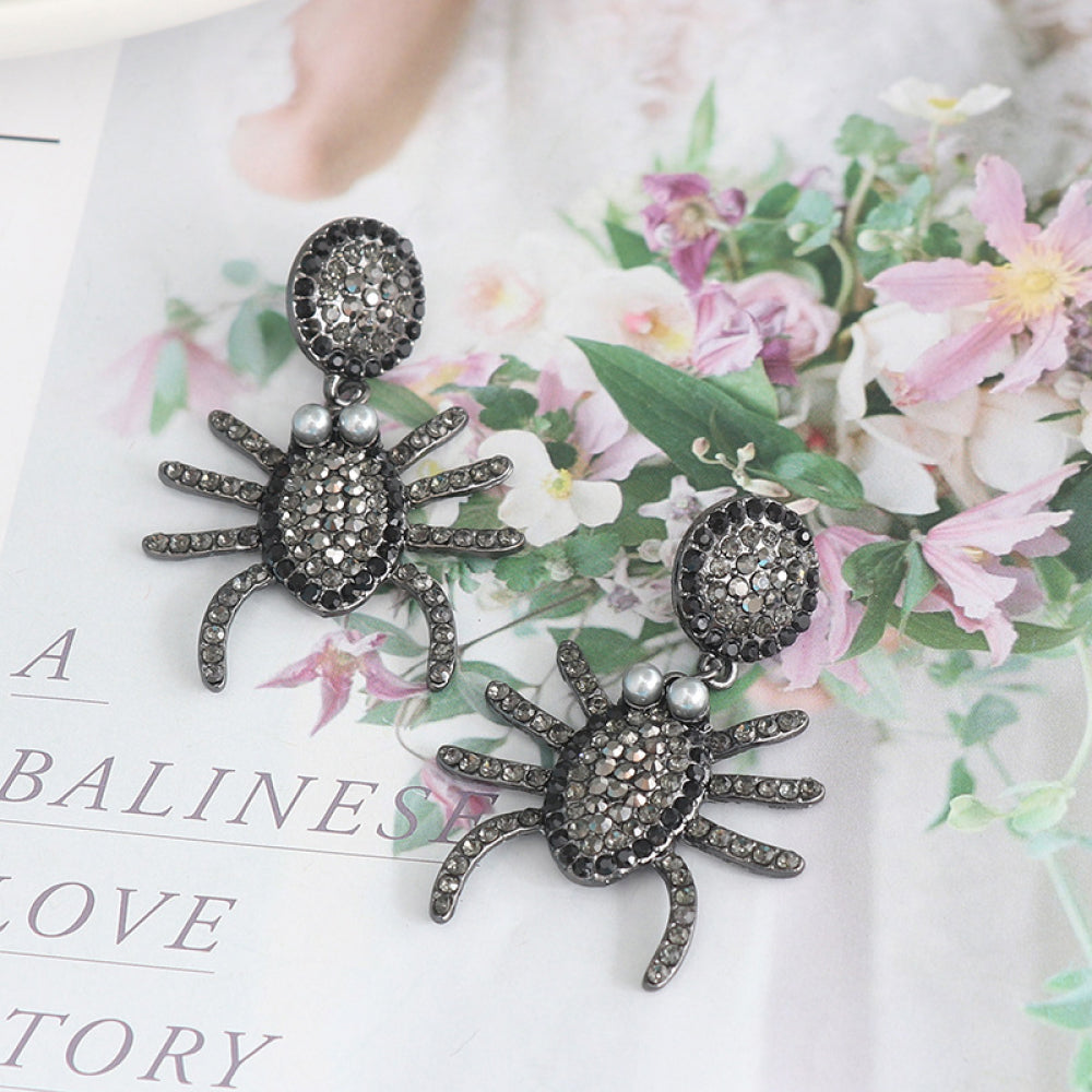 Two dark gray rhinestone spider earrings with pearl accents lie on a floral background.