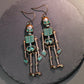 Two antique bronze skeleton alloy earrings with teal accents hang on a dark surface.