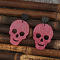 Two pink glitter skull earrings with black heart studs are displayed on bamboo sticks.