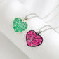 Two heart-shaped necklaces, one green and one pink, with spiderweb designs and glitter.