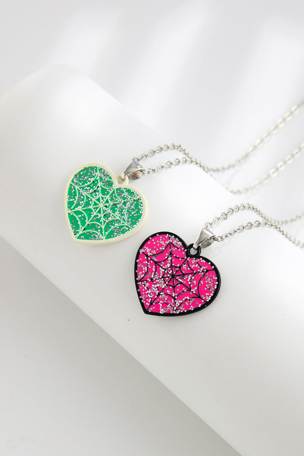 Two heart-shaped necklaces, one green and one pink, with spiderweb designs and glitter.
