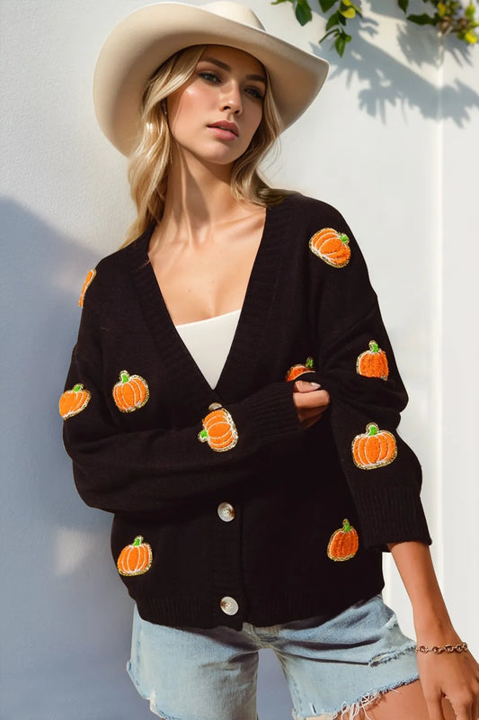 A woman wears a black button-up cardigan with pumpkin appliques and light wash shorts.