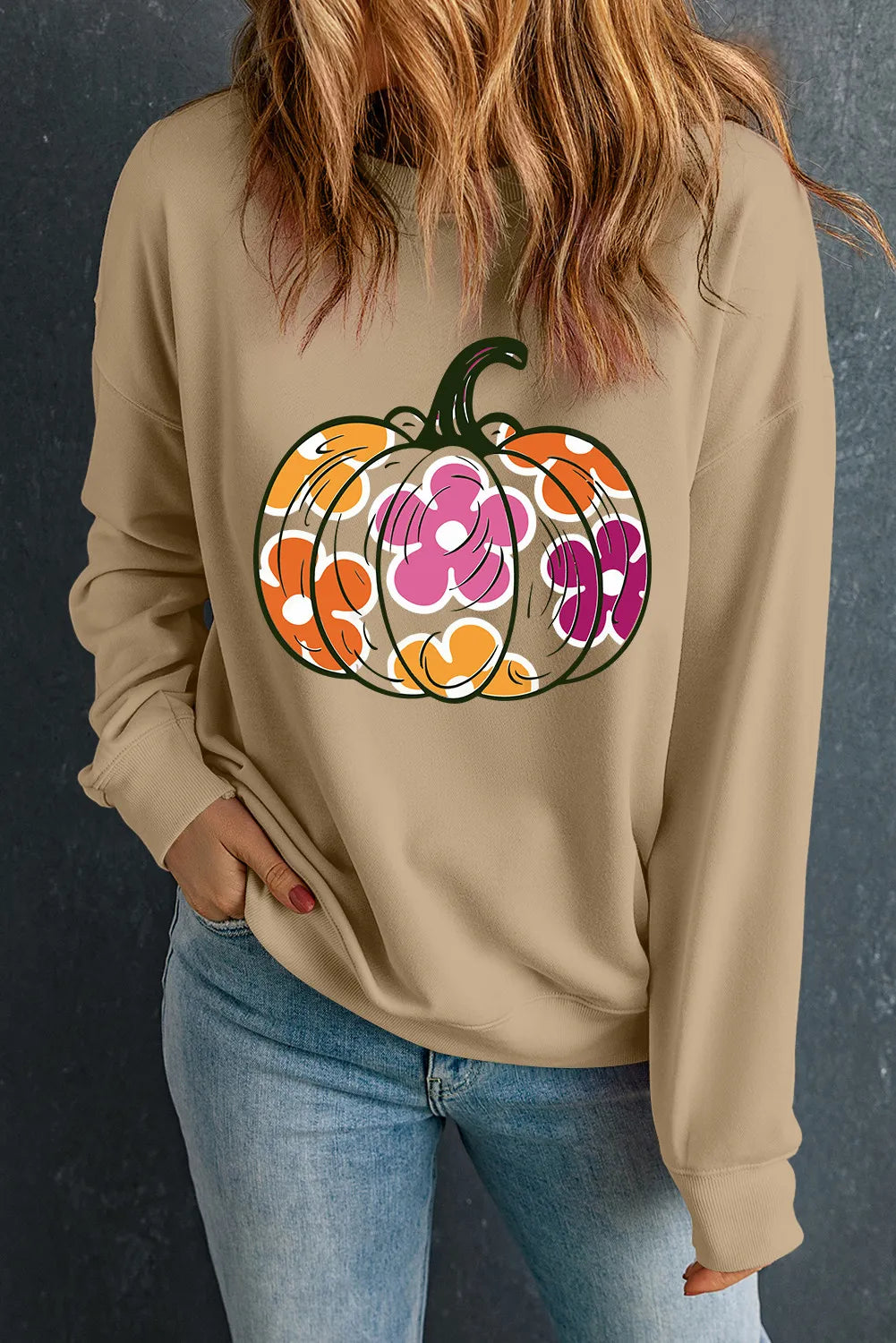 A woman wears a tan long-sleeve sweatshirt with a colorful pumpkin and flower graphic.