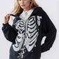 A woman wears a black skeleton zip-up drawstring hoodie over a white top and lace skirt.