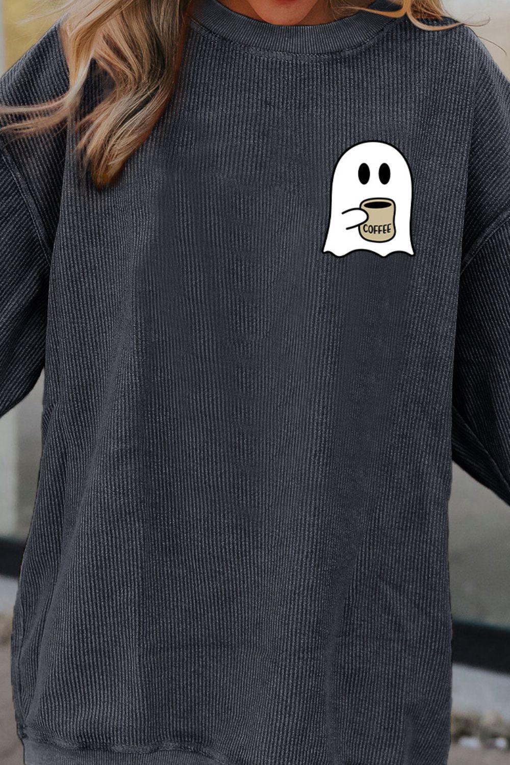 A woman wears a dark gray drop-shoulder sweatshirt with a ghost graphic holding coffee.