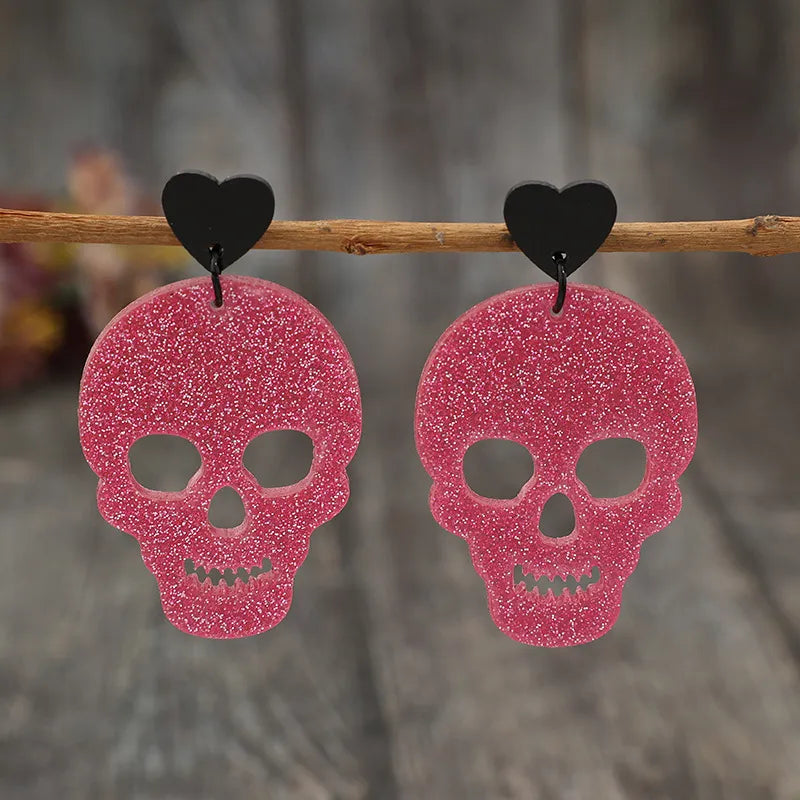 Two glittery pink skull earrings with black heart studs hang from a wooden branch.