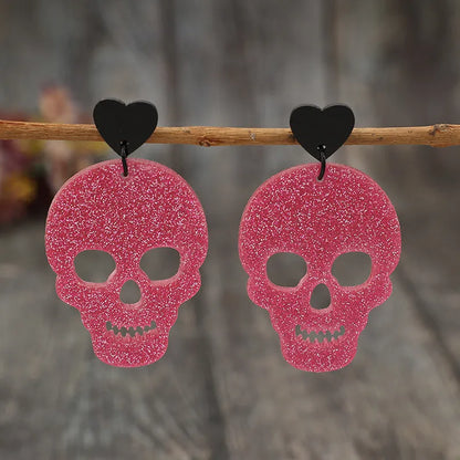 Two glittery pink skull earrings with black heart studs hang from a wooden branch.
