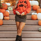 A woman wears an orange pumpkin pattern round neck sweater with a dark green skirt and black boots.