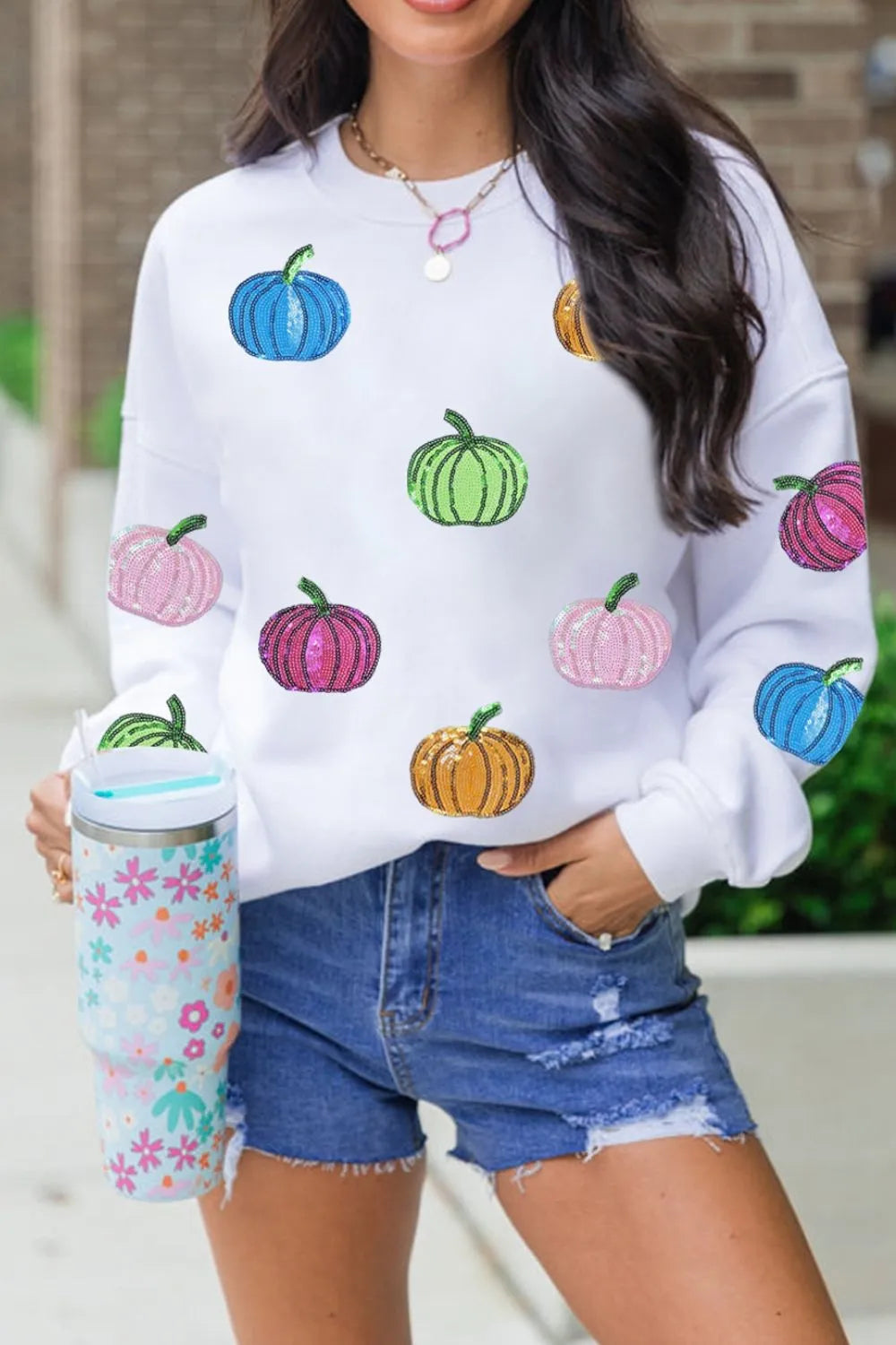 White long-sleeve sweatshirt with colorful sequin pumpkins and distressed denim shorts.